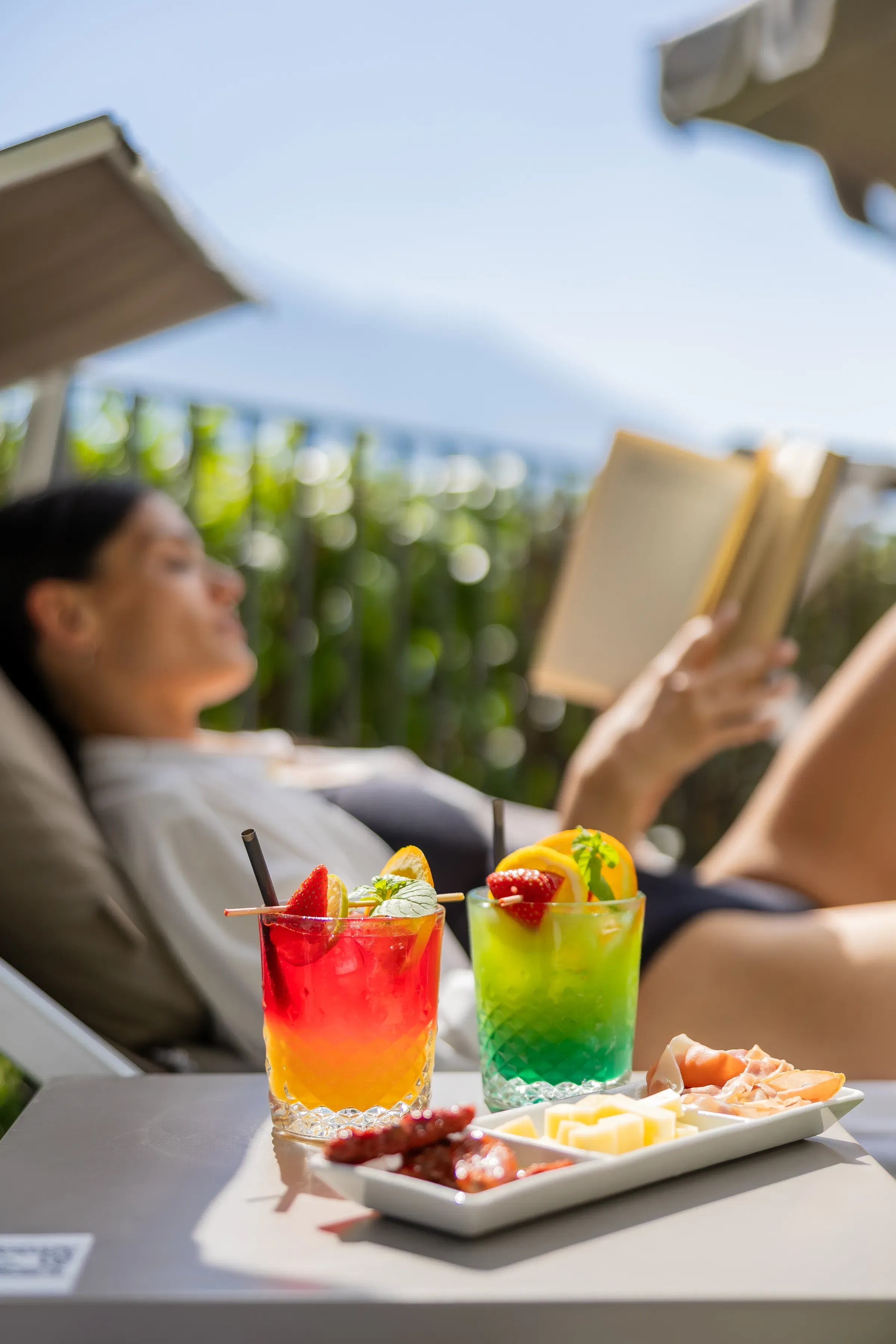 Cocktail colorati e stuzzichini su tavolino con donna che legge sullo sfondo