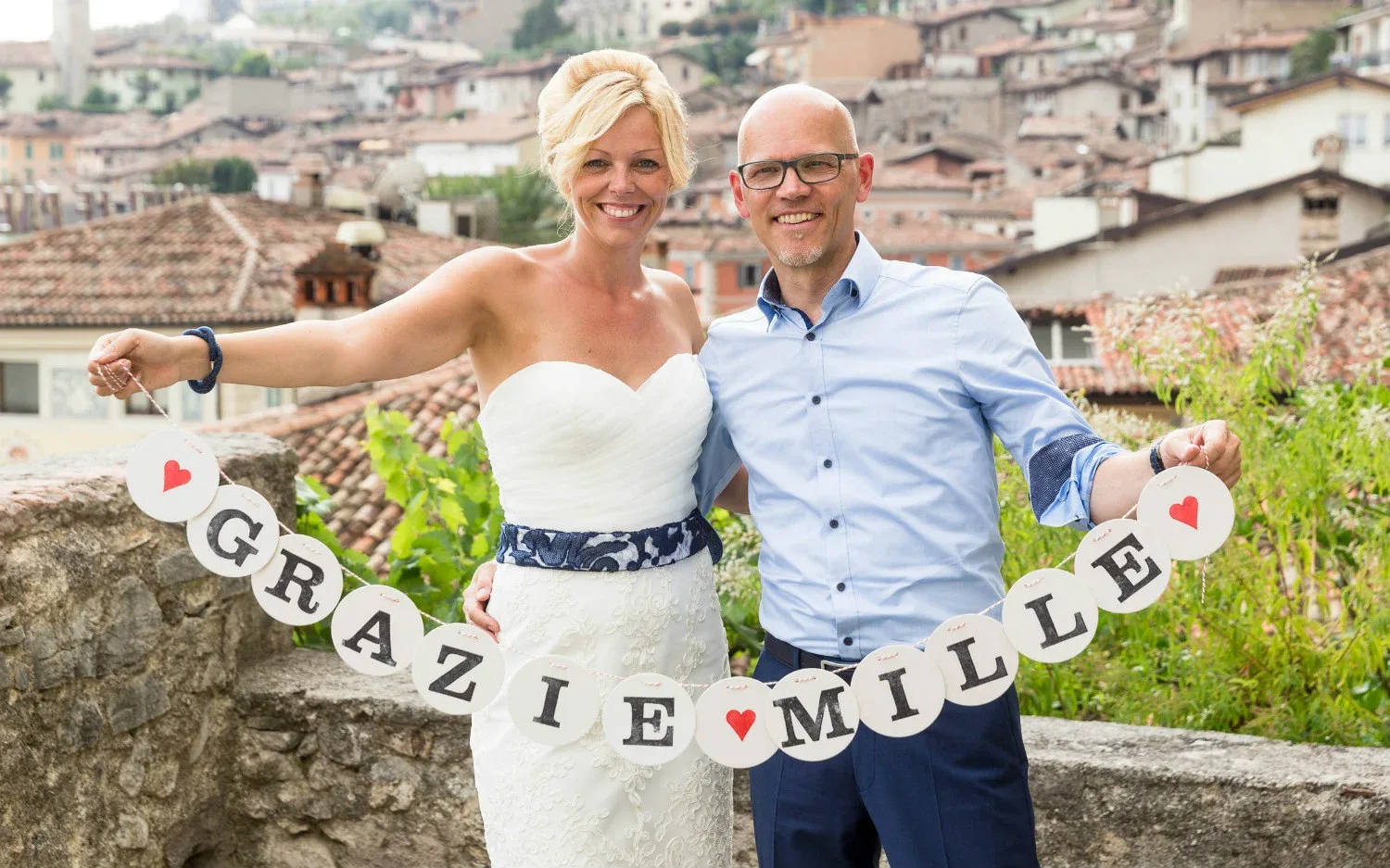 Lächelndes Hochzeitspaar mit 'Danke'-Banner in einer italienischen Stadt